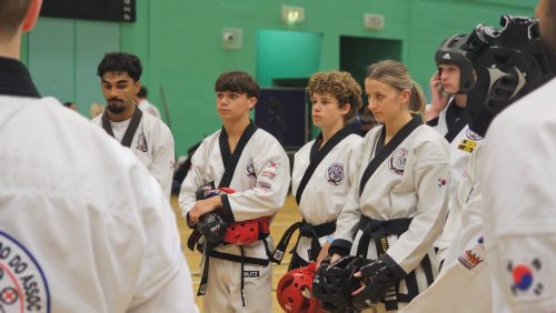 Black belt students standing in line with sparring gear, watching a demonstration at the club's August 2026 clinic